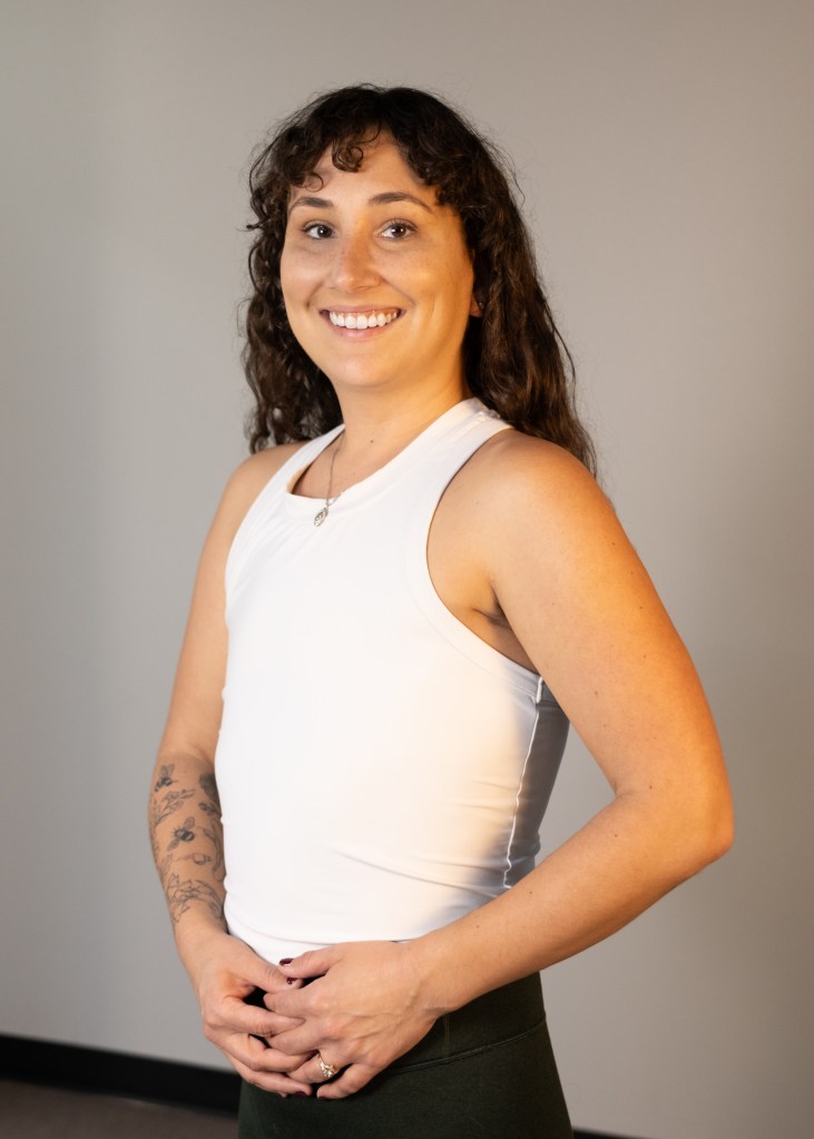 A smiling yoga and meditation teacher poses confidently in a white tank top and green pants, standing against a neutral background.